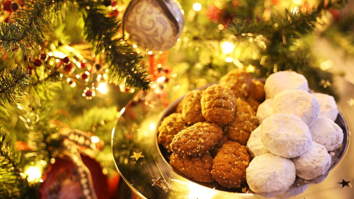 A festive Christmas setting with a close-up of a plate filled with traditional Greek Christmas cookies, melomakarona and kourabiedes, in front of a beautifully decorated Christmas tree adorned with lights and ornaments.